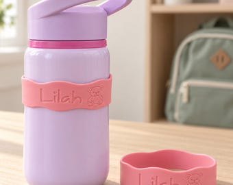 Può includere: Una borraccia color viola chiaro con manico e una fascia in silicone rosa che riporta il nome Lilah e una piccola icona di orsetto. Una fascia in silicone rosa abbinata con lo stesso nome e disegno di orsetto è posizionata accanto alla borraccia.