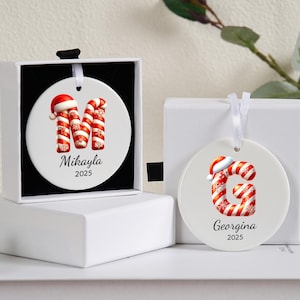 Puede incluir: Dos adornos navideños redondos de cerámica blanca. Uno presenta la letra "M" hecha de bastones de caramelo rojos y blancos, un gorro de Papá Noel, el nombre "Mikayla" y el año "2025". El otro presenta la letra "G", el nombre "Georgina" y el año "2025".