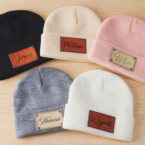 May include: Five knit beanies in different colors with personalized leather patches. The patches have the names "Dorcie", "Willow", "Hallie", "Mcwhish", and "Wyatt" written on them.