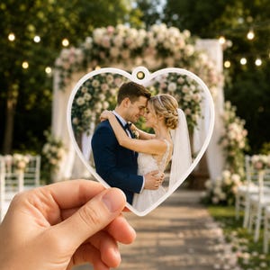 May include: A heart-shaped ornament featuring a printed photograph of a couple embracing at their wedding. The ornament has a white frame and a small hole at the top for hanging. The couple is dressed in formal wedding attire in an outdoor setting.