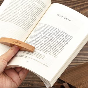 May include: A wooden book page holder with the name "Sophia" engraved on it, resting on an open book. The holder is light brown and designed to slip over a finger for easy reading. The book is open to Chapter IV.