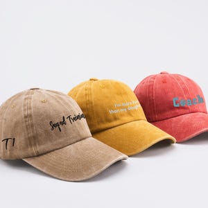 May include: Three baseball caps with different embroidered text. The first cap is brown with the text "Squad Training" and "T1". The second cap is yellow with the text "I'm more fun than my daughter". The third cap is red with the text "Coach" in blue.