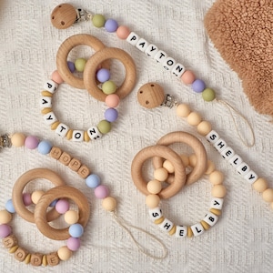 May include: Wooden baby pacifier clips and teething rings. The clips have a wooden design with a metal clasp. The teething rings are made of wood and colourful silicone beads. The names "PAYTON", "HOPE", and "SHELBY" are spelled out on the clips.