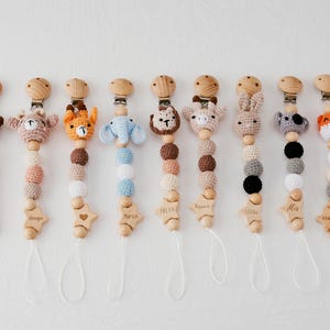 May include: A collection of baby pacifier clips with wooden beads and crochet animal heads. Each clip features a different animal, including a bear, giraffe, elephant, and fox. The clips have personalized wooden star-shaped name tags.