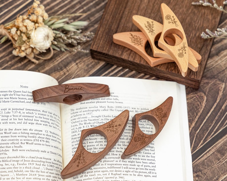 May include: Wooden thumb page holders in various shades of brown, some with floral engravings. One holder is engraved with the name "Bonnie". These accessories are designed to hold open a book.