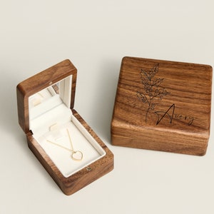 Pu&ograve; includere: Una scatola di legno con un coperchio a cerniera, incisa con un motivo floreale e il nome "Avery". La scatola &egrave; aperta, rivelando una piccola collana a forma di cuore all'interno.