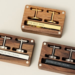 Puede incluir: Tres cajas de madera con gemelos y pasadores de corbata grabados. Las cajas son de madera oscura y tienen un acabado pulido. Los gemelos y los pasadores de corbata son de plata y oro. Las cajas están etiquetadas como "The Best Man", "Father of the Groom" y "Father of the Groom".