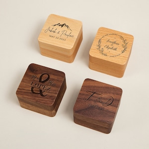 May include: Four square wooden ring boxes. Two are light-coloured with engraved names and dates, one with a mountain design. The other two are dark wood, one with "Mr & Mrs Lopez", the other with initials and a date.