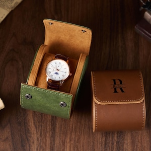 May include: A green leather watch case with a brown interior holds a gold-toned watch with a white face and brown leather band. A closed brown leather watch case with the letter "R" and the name "Rachel" is also present.