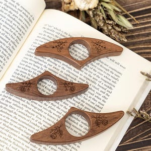 May include: Three wooden thumb page holders with floral engravings rest on an open book. The page holders are a warm brown color and have a circular opening for the thumb. The book is open to a page of text.