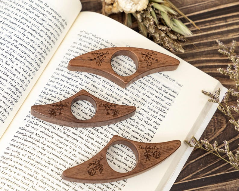 May include: Three wooden thumb page holders with floral engravings rest on an open book. The page holders are a warm brown color and have a circular opening for the thumb. The book is open to a page of text.