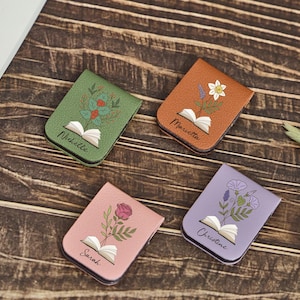 May include: Four rectangular magnetic bookmarks in green, tan, pink, and lavender. Each features an open book illustration with a different flower growing from the pages and a printed name: Nichelle, Marietta, Sarah, and Christine.