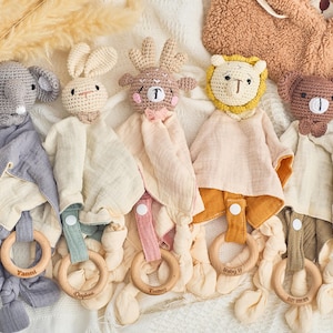 K&ouml;nnte beinhalten: F&uuml;nf geh&auml;kelte Babyspielzeuge mit befestigten Musselin-Decken. Die Spielzeuge sind ein grauer Elefant, ein braunes Reh, ein brauner B&auml;r, ein wei&szlig;es Kaninchen und ein gelber L&ouml;we. Jedes Spielzeug hat einen Holzring, der an der Decke befestigt ist.