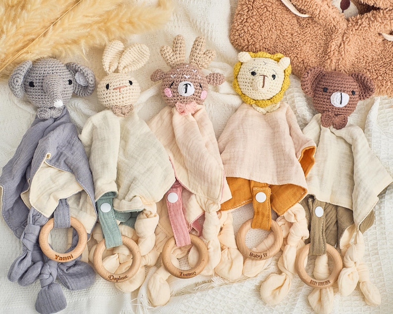 K&ouml;nnte beinhalten: F&uuml;nf geh&auml;kelte Babyspielzeuge mit befestigten Musselin-Decken. Die Spielzeuge sind ein grauer Elefant, ein braunes Reh, ein brauner B&auml;r, ein wei&szlig;es Kaninchen und ein gelber L&ouml;we. Jedes Spielzeug hat einen Holzring, der an der Decke befestigt ist.
