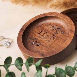 May include: A wooden ring dish with a floral design and the initials "H&B" engraved on it. The date "08-27-2023" is also engraved on the dish.