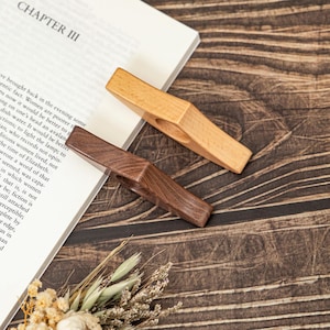 May include: Two wooden book page holders, one light-colored and one dark brown, rest on a book with the title "CHAPTER III" visible. The holders are angled, showcasing their simple, functional design. The background features a wooden surface and dried flowers.