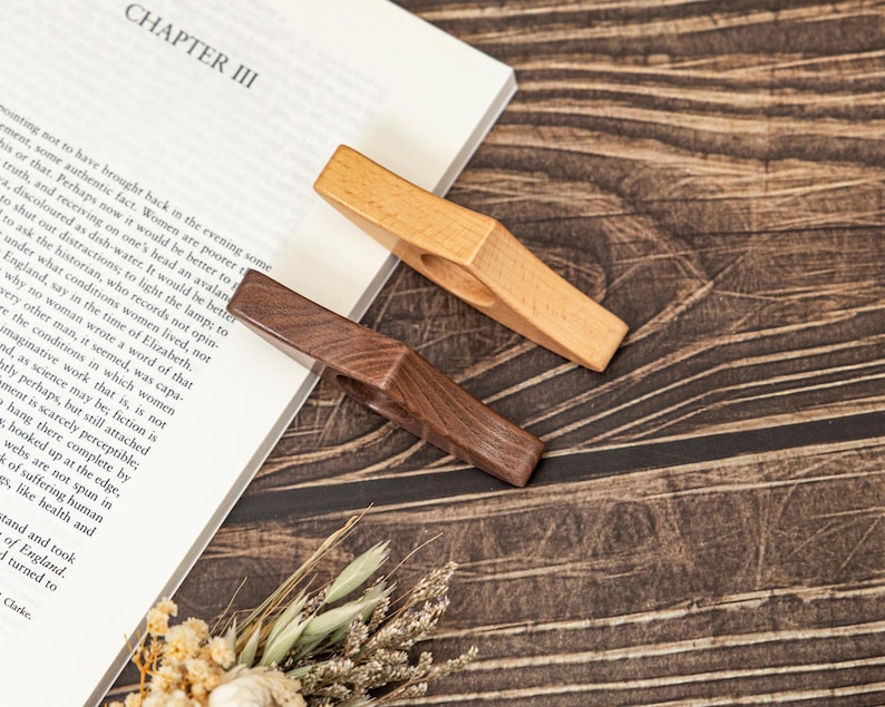 May include: Two wooden book page holders, one light-colored and one dark brown, rest on a book with the title "CHAPTER III" visible. The holders are angled, showcasing their simple, functional design. The background features a wooden surface and dried flowers.