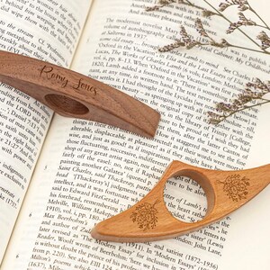 May include: Two wooden thumb page holders, one with the name "Romy Jones" engraved, and the other with a floral design. The page holders are resting on an open book.