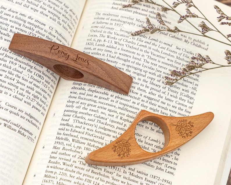 May include: Two wooden thumb page holders, one with the name "Romy Jones" engraved, and the other with a floral design. The page holders are resting on an open book.