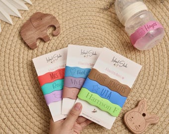 May include: A collection of baby accessories, including personalized silicone bands with names and shapes, a baby bottle with a pink band, and wooden toys. The bands are in various colors, with names like "Mrs R" and "Harrison F".