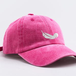 May include: A pink cotton baseball cap with a white feather design embroidered on the front.