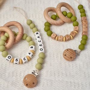 May include: Baby pacifier clips and teething rings in olive green and natural wood. The pacifier clips feature the names "Oscar" and "Viaan" spelled out with lettered beads. The wooden rings are smooth and round.