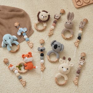 May include: A collection of handmade crochet baby rattles and pacifier clips featuring various animal designs including a lion, elephant, rabbit, koala, fox, and giraffe. Each item incorporates wooden rings, beads, and metal clips with names engraved.