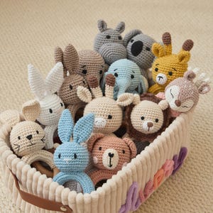 May include: A collection of crocheted animal rattles in a cream-colored corduroy basket. The set includes a white rabbit, a blue rabbit, a brown bear, a lion, a giraffe, an elephant, a koala, a hippo, and a deer, all with stitched features.