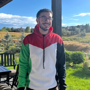 May include: A color-blocked hoodie with a red, white, green, and black design. The hoodie has a full zipper and a hood. The person is wearing brown pants. The background includes a green lawn, trees, and a blue sky.