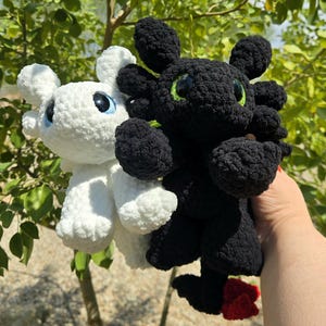 May include: Two crocheted dragon plushies. One is white with blue eyes, the other is black with green eyes. Both have large ears and small red accents. The plushies are handmade and appear soft.
