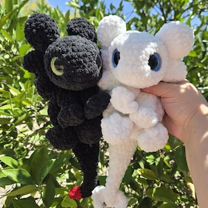 Puede incluir: Dos peluches de drag&oacute;n hechos a mano con ganchillo, uno negro y otro blanco, se sostienen contra un fondo de hojas verdes y cielo azul. El drag&oacute;n negro tiene ojos verdes y una punta de cola roja. El drag&oacute;n blanco tiene ojos azules.