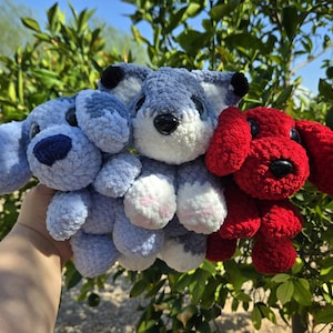 May include: Three handmade crochet dog stuffed animals in blue, gray, and red. The blue dog has a dark blue nose, the gray dog has black ears, and the red dog has floppy ears. The dogs are held in front of green foliage.