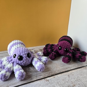 May include: Two crocheted octopus plush toys. One is lavender with white accents, the other is dark purple with black accents. Both have large black eyes and are displayed on a wooden surface.