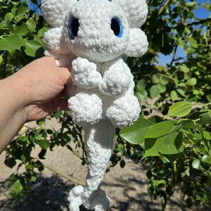 Puede incluir: Un peluche blanco hecho a mano, que se asemeja a un drag&oacute;n o criatura m&iacute;tica. Tiene ojos azules grandes y redondos y detalles esponjosos y texturizados. El juguete se sostiene contra un fondo de follaje verde y cielo azul.