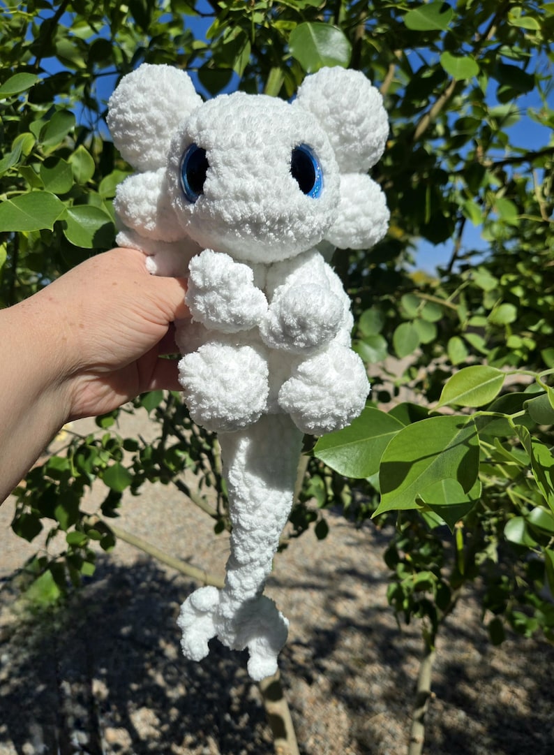 Puede incluir: Un peluche blanco hecho a mano, que se asemeja a un drag&oacute;n o criatura m&iacute;tica. Tiene ojos azules grandes y redondos y detalles esponjosos y texturizados. El juguete se sostiene contra un fondo de follaje verde y cielo azul.