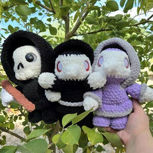 May include: Three crocheted figures: a Grim Reaper with a scythe, a black-robed figure, and a purple-clad figure. The Grim Reaper has a skull face and black hood. The other figures have white faces and red eyes. The figures are handmade.