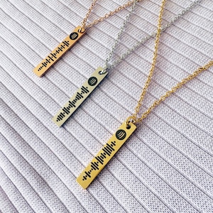 May include: Three necklaces with rectangular pendants. One is rose gold, one silver, and one gold. Each pendant displays a black graphic, possibly a sound wave or barcode, suspended from fine chains. The necklaces are displayed on a textured surface.