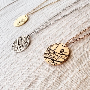 Peut inclure: Trois colliers avec des pendentifs ovales représentant des cartes. Les chaînes sont dorées, argentées et or rose. Les pendentifs présentent des dessins de cartes en noir. Les colliers sont présentés sur une surface texturée blanc cassé.