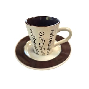 May include: A ceramic coffee cup and matching saucer set. The cup is cream-colored with a dark brown interior and handle, featuring vertical text that reads coffee and a column of brown coffee bean illustrations. The saucer has a dark brown rim.