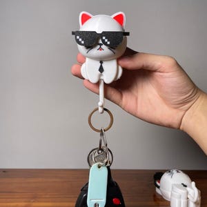 Puede incluir: Un llavero blanco con forma de gato con detalles de orejas rojas y gafas de sol pixeladas. El gato tiene una corbata negra y un llavero adjunto. Una mano sostiene el llavero, con llaves y una etiqueta azul adjuntas.