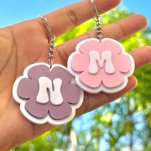 May include: Two flower-shaped keychains, one lavender and one pink, each with a white letter. The lavender keychain has the letter "N", and the pink keychain has the letter "M". Both keychains have silver key rings.