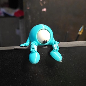 May include: A light blue, 3D-printed desk accessory shaped like a cartoon character with a large white eye and black pupil. The character has small arms and legs, and is perched on the edge of a black laptop.
