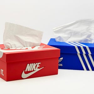 Puede incluir: Cajas de pañuelos de papel rojas y azules diseñadas para parecerse a cajas de zapatos Nike y Adidas. La caja roja tiene el logotipo de Nike y el texto "JUST DO IT". La caja azul presenta el logotipo y las rayas de Adidas. Cada caja dispensa pañuelos blancos.