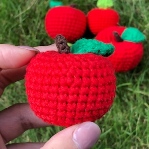 Pode incluir: Quatro maçãs vermelhas de crochê com folhas verdes. As maçãs estão em uma mão, e o fundo é grama verde.