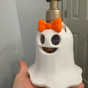 May include: A white ghost-shaped soap dispenser holder featuring a bright orange bow. The holder is designed to fit around a standard foaming hand soap bottle with a gold-colored pump dispenser. The ghost has two eye cutouts and a mouth cutout.