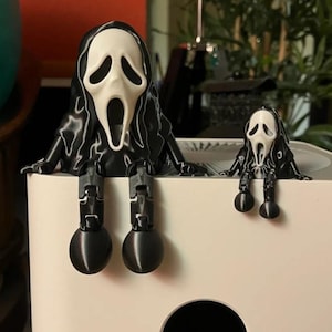May include: Two Ghostface figures with white masks and black hooded robes are perched on the edge of a white surface. Each figure features articulated black limbs with rounded feet, mimicking a seated position with legs dangling over the edge.