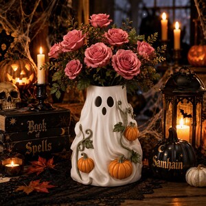 May include: A white ceramic ghost-shaped vase featuring a cutout face and sculpted orange pumpkins with green vines. The vase is filled with a bouquet of pink roses and green foliage, creating a decorative piece for seasonal displays.