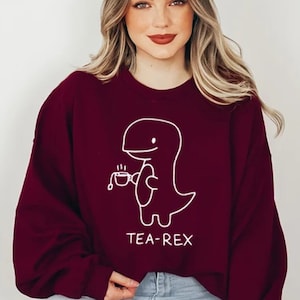 May include: A maroon sweatshirt with a white graphic of a dinosaur holding a cup of tea. The text "TEA-REX" is printed below the dinosaur.