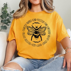 May include: A mustard yellow short-sleeve t-shirt featuring a black graphic of a bee centered within a circular border. The text surrounding the bee includes the words: HUMBLE, KIND, POSITIVE, FUN, CONFIDENT, BRAVE, SMART, HAPPY, WILD, CREATIVE, JOYFUL, YOURSELF.
