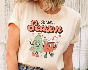 May include: A beige t-shirt with a retro-style graphic of a Christmas tree and a mug of hot chocolate with the text "Tis' the Season".